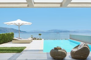Zona de piscina con sillones de descanso y una sombrilla, con vista a un cuerpo de agua y montañas a lo lejos.