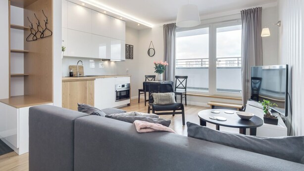 Een moderne woonkamer met een grijze bank, een eettafel met stoelen, een televisie en een keuken met witte kasten en een houten aanrecht.
