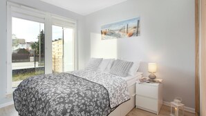 Een slaapkamer met een bed, nachtkastje, lamp, schilderij en uitzicht door een raam.
