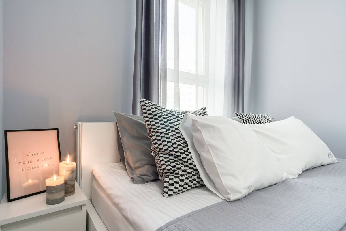 Une chambre à coucher bien rangée, avec un lit, des oreillers, une table de chevet avec des bougies et un cadre contenant une citation.