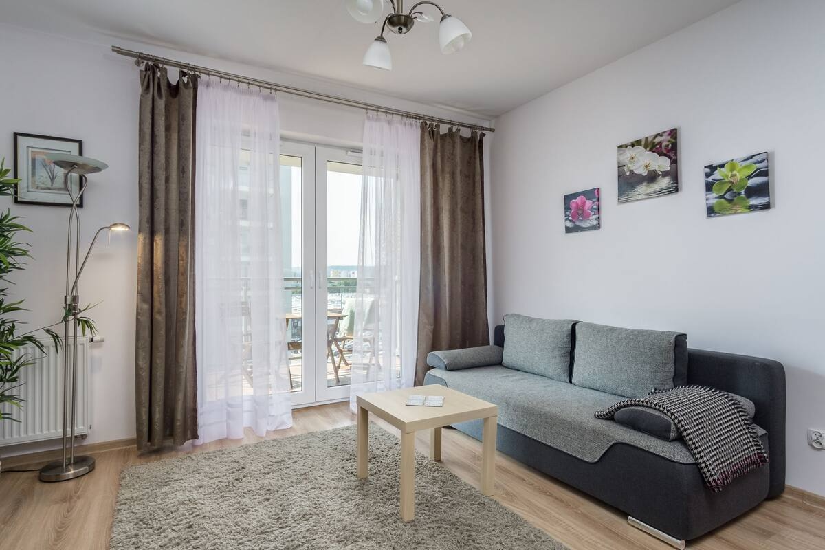 Un salon moderne avec un canapé gris, une petite table en bois et une vue sur un espace extérieur, visible à travers une porte vitrée.