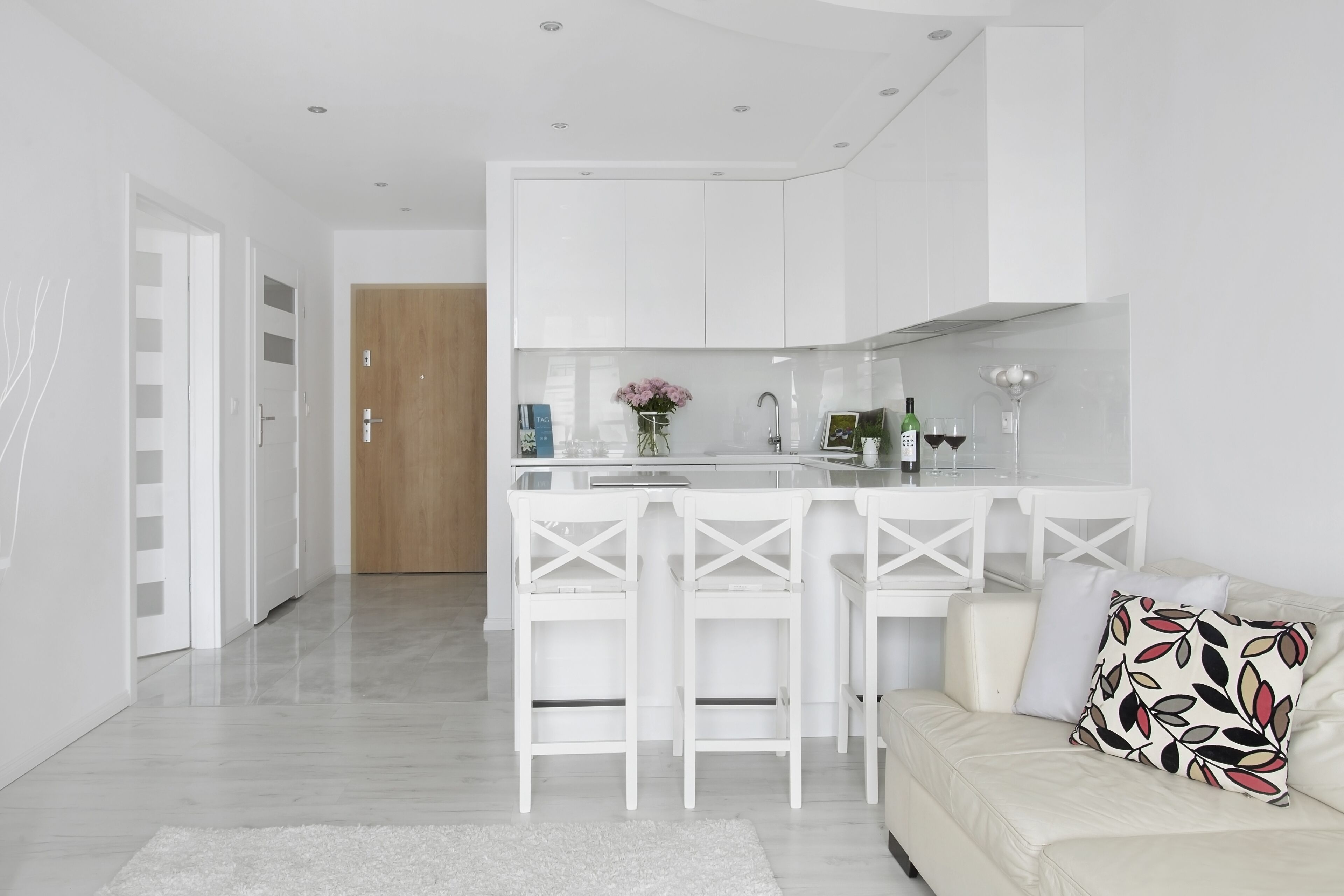 Una cocina moderna con armarios blancos, una isla central con taburetes y un sofá blanco con un cojín estampado.