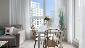 A modern living room with a white sofa, a dining table set with chairs, and a large window with a city view. - Elite Apartments Marine Cztery Oceany (Gdansk)