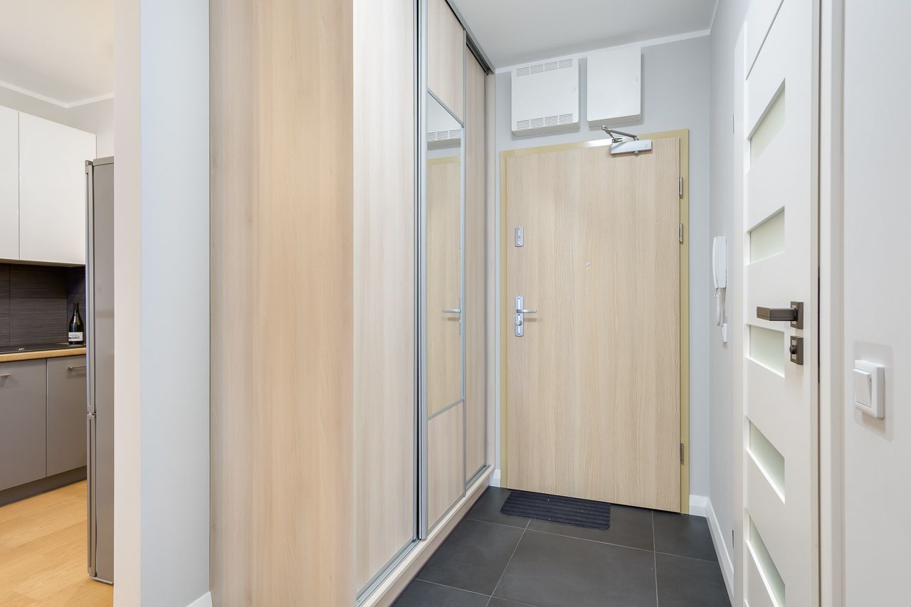 En modern hall med en trädörr, ett köksområde med ett kylskåp och en vit vägg med en väggmonterad luftkonditioneringsanläggning.