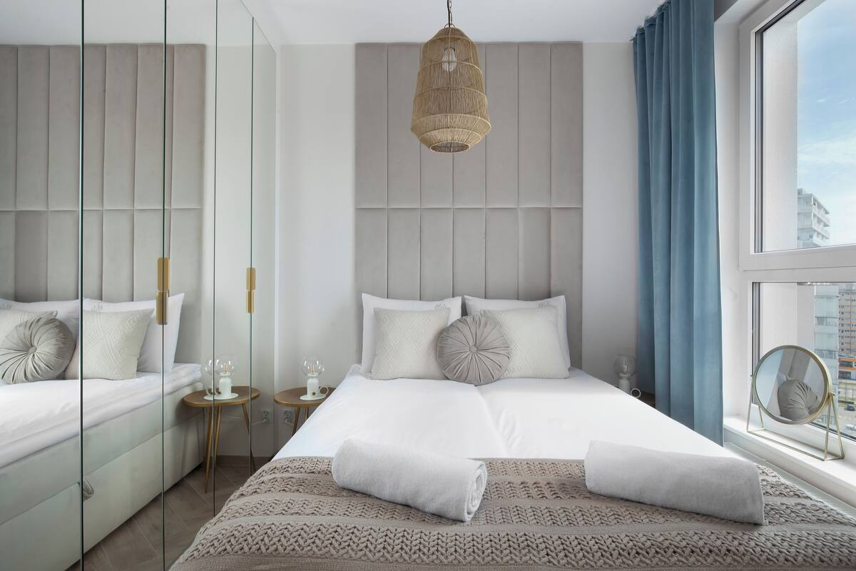 Une chambre d’hôtel moderne avec un grand lit, une table de chevet, un luminaire suspendu et une vue sur la ville par la fenêtre.