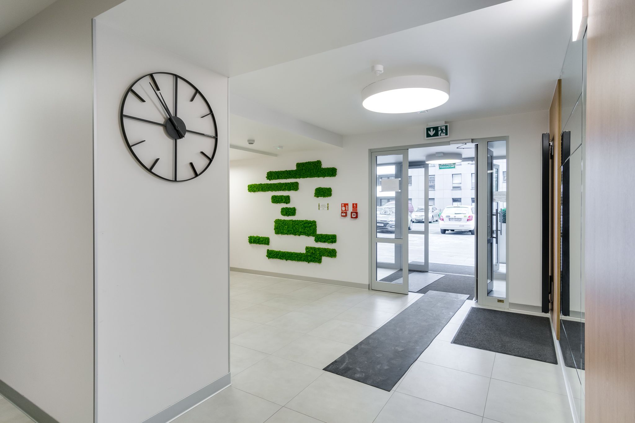 A modern lobby with a large wall clock, a green wall art installation, and a door leading outside.