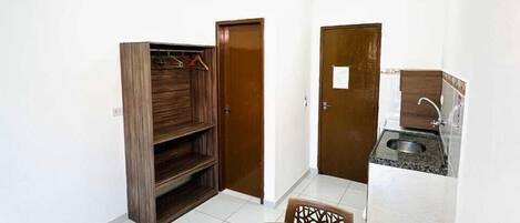 Una habitación pequeña y limpia con una mesa redonda de madera y dos sillas, un armario de madera, una puerta y una kitchenette con un fregadero y encimera.