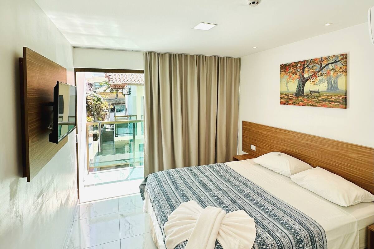 A hotel room with a bed, a painting, a TV, and a view of the outside.