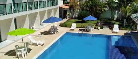 A swimming pool with lounge chairs and umbrellas, surrounded by a building with large windows and a view of palm trees.