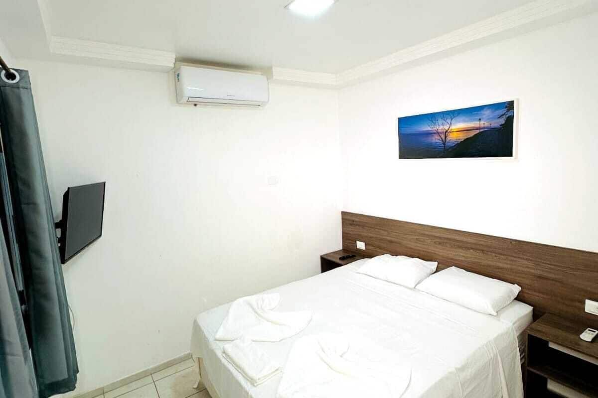 A hotel room with a bed, a nightstand, a wall-mounted TV, an air conditioning unit, and a painting of a sunset.