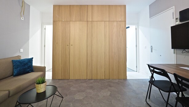 Una sala de estar moderna con un sofá, una mesa de centro, una mesa de comedor con sillas, un televisor montado en la pared y un amplio armario de madera.
