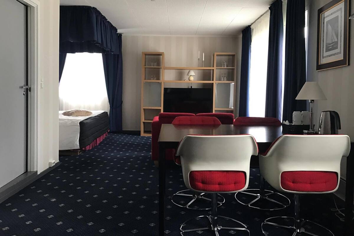 A hotel room with a bed, a TV, a dining area with a table and chairs, a bookshelf, and a window with curtains.