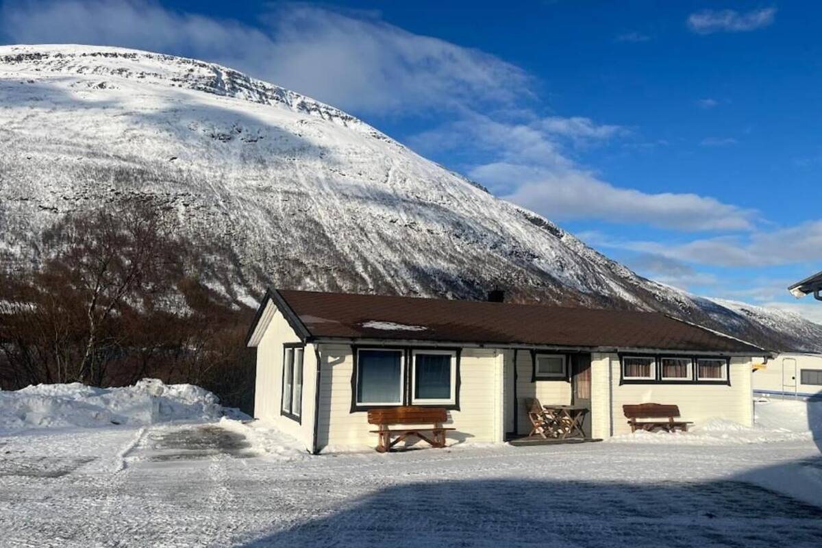 A small, single-story building with a sloped roof, a front porch with benches, and a snow-covered landscape with a mountain in the background.