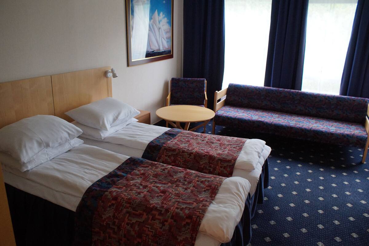 A hotel room with two beds, a small round table, two patterned armchairs, and a painting on the wall.