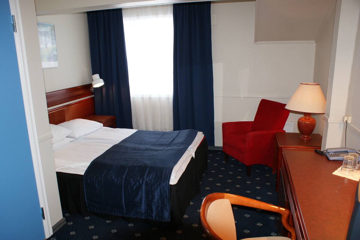 A hotel room with a bed, a desk, a chair, a lamp, and a window with curtains.