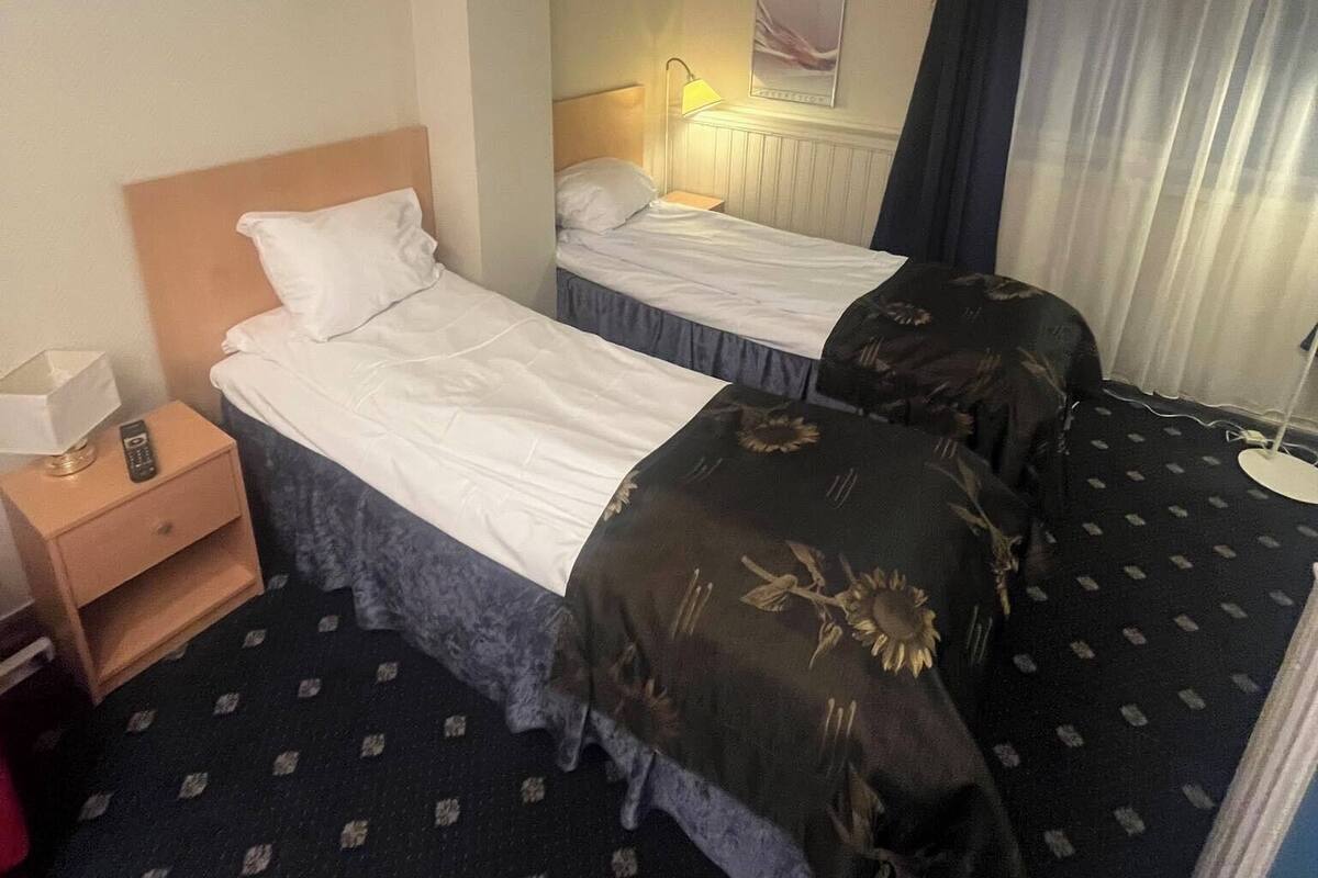 A hotel room with two beds, a bedside table with a lamp, a mirror, and a window with curtains.