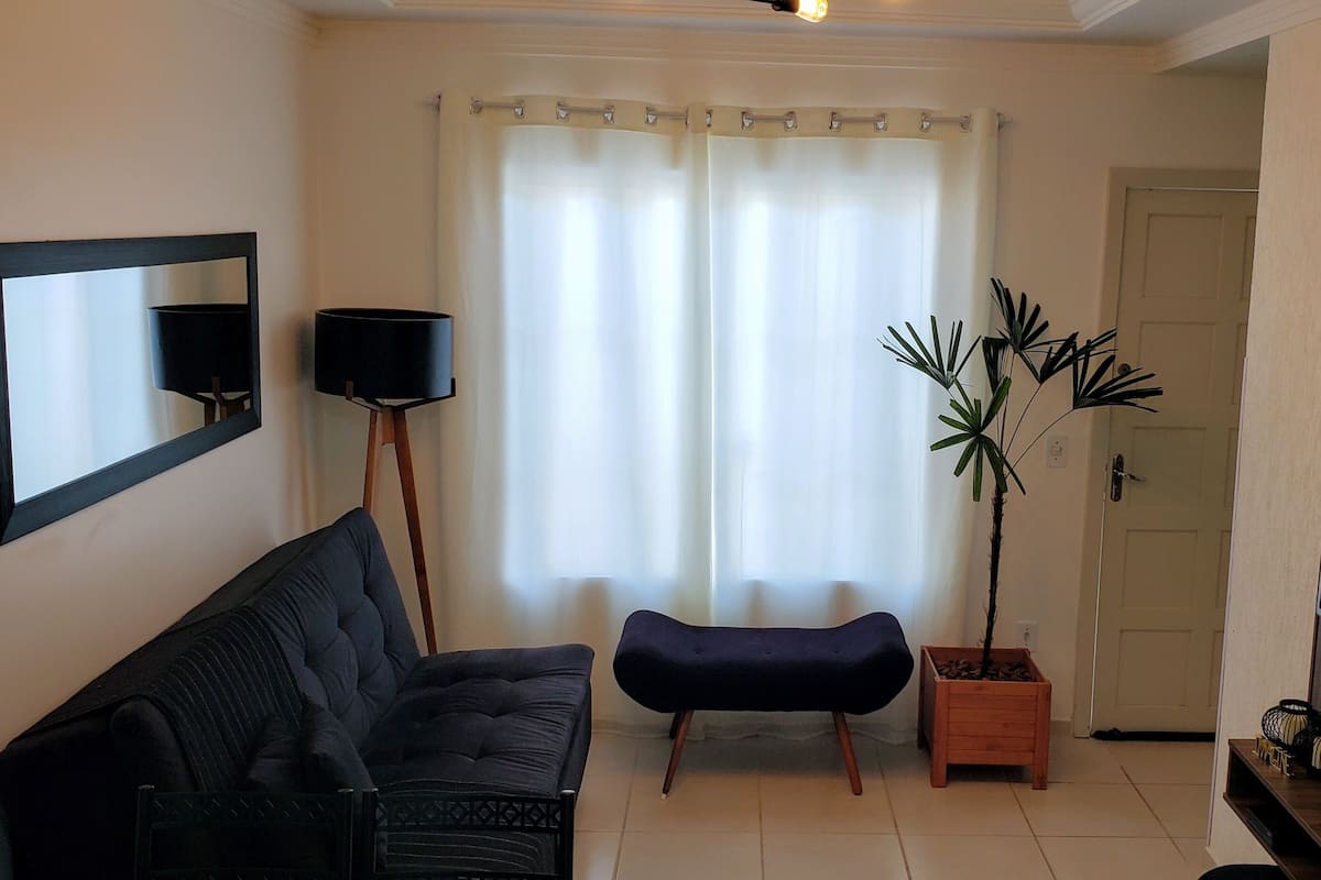 A living room with a black sofa, a small ottoman, a floor lamp, a potted plant, and a flat-screen TV mounted on the wall.