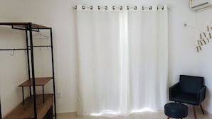 A room with a white curtain, a black chair, a wooden shelf unit, and a wall-mounted air conditioner. - Residencial Catarina (Florianópolis)