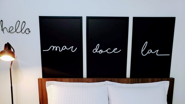 A bed with white bedding and two pillows, a bedside lamp, and three black wall art pieces with white cursive text. - Residencial Catarina (Florianópolis)