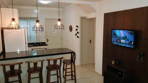 A modern kitchen with a central island, bar stools, pendant lighting, and a flat-screen TV mounted on the wall. - Residencial Catarina (Florianópolis)