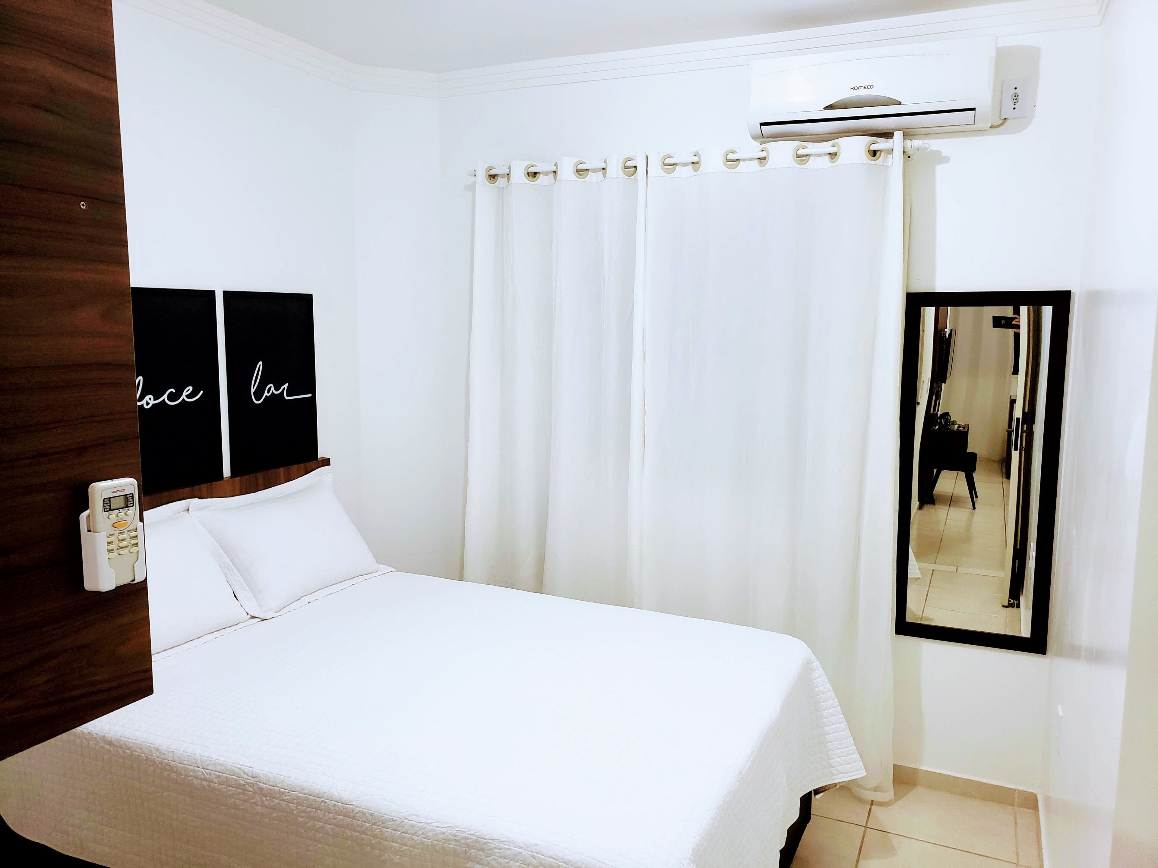 A hotel room with a bed, white curtains, a mounted air conditioner, a bedside phone, and a mirror on the wall.
