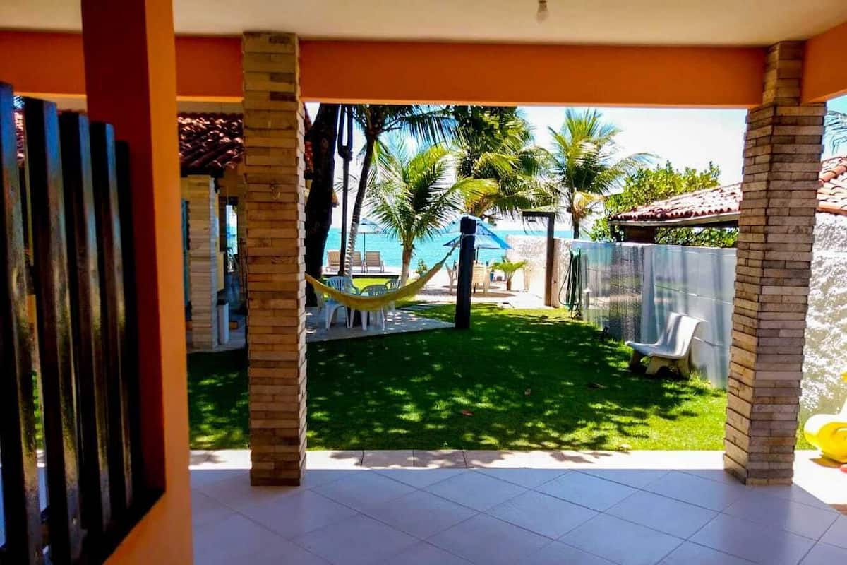 Uma área de varanda coberta com piso ladrilhado, uma rede e vista para uma piscina e palmeiras.