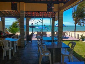 A covered outdoor seating area with tables and chairs, overlooking a beach with umbrellas and palm trees. - Pousada Santo Aleixo (Ipojuca)