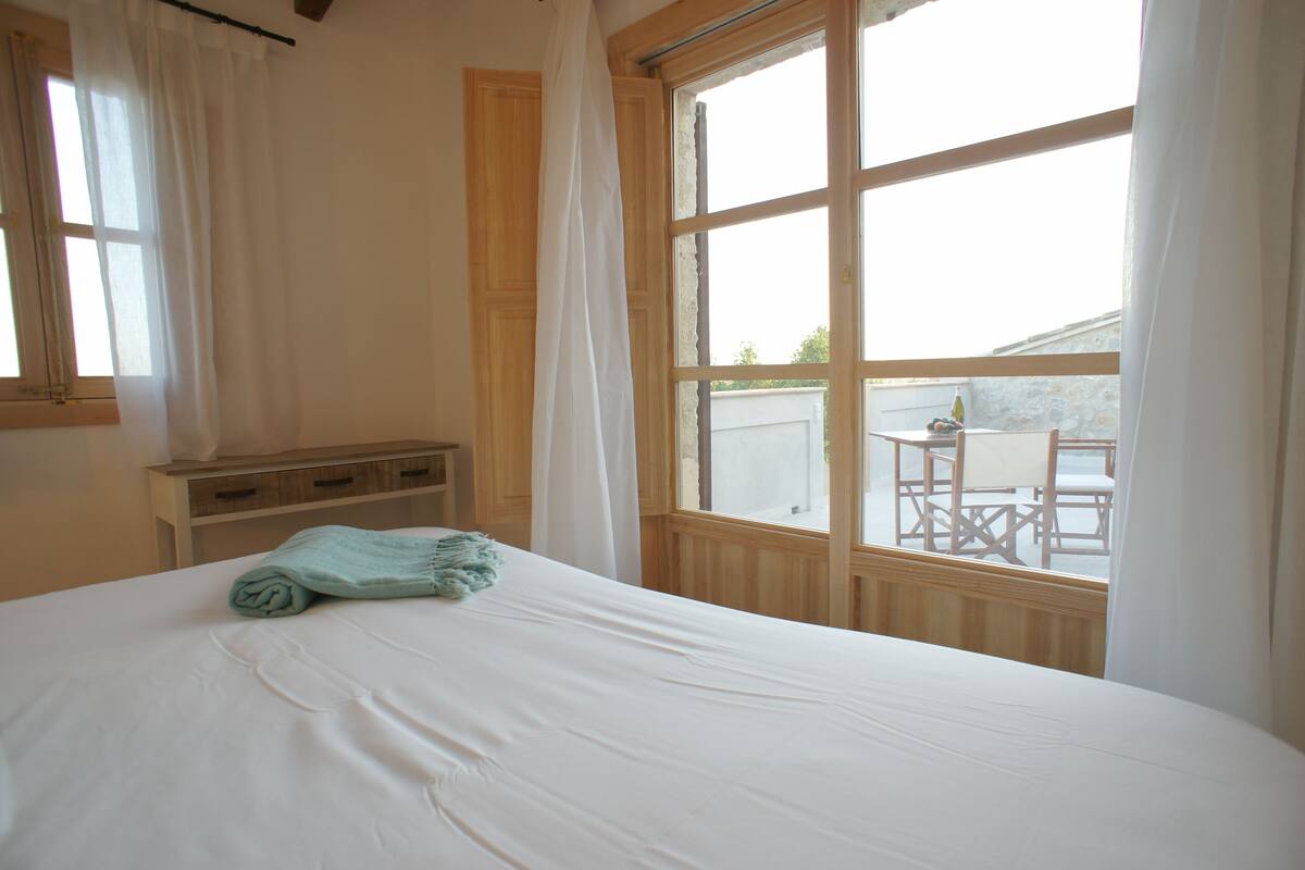 A bedroom with a large window, a bed, a wooden nightstand, and a view of an outdoor patio.