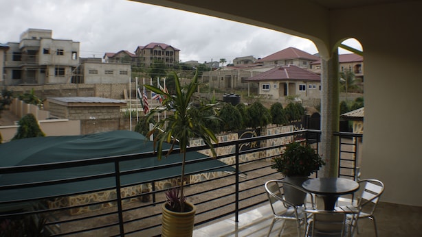 A balcony with a green table and chairs, a potted plant, and a view of residential buildings. - Dokua Royal Hotel (Kwabenya)