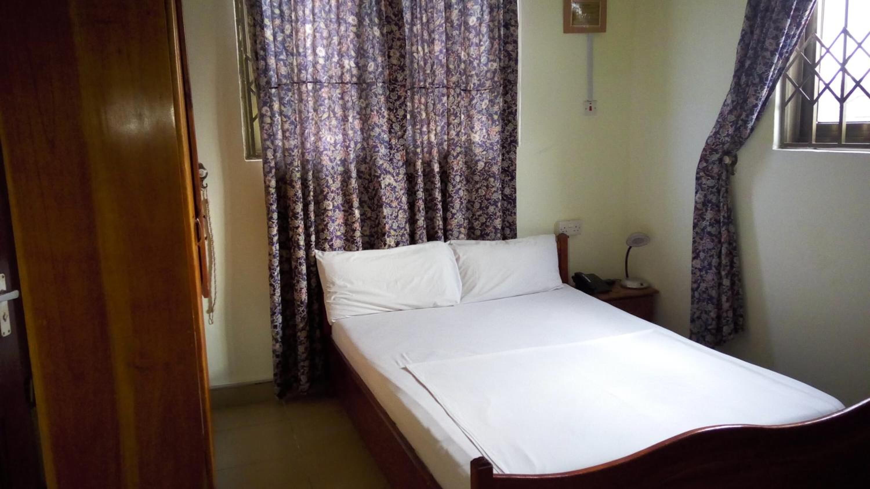 Una cama individual con ropa de cama blanca en una habitación con cortinas florales, una mesita de noche con lámpara y un aplique de pared.