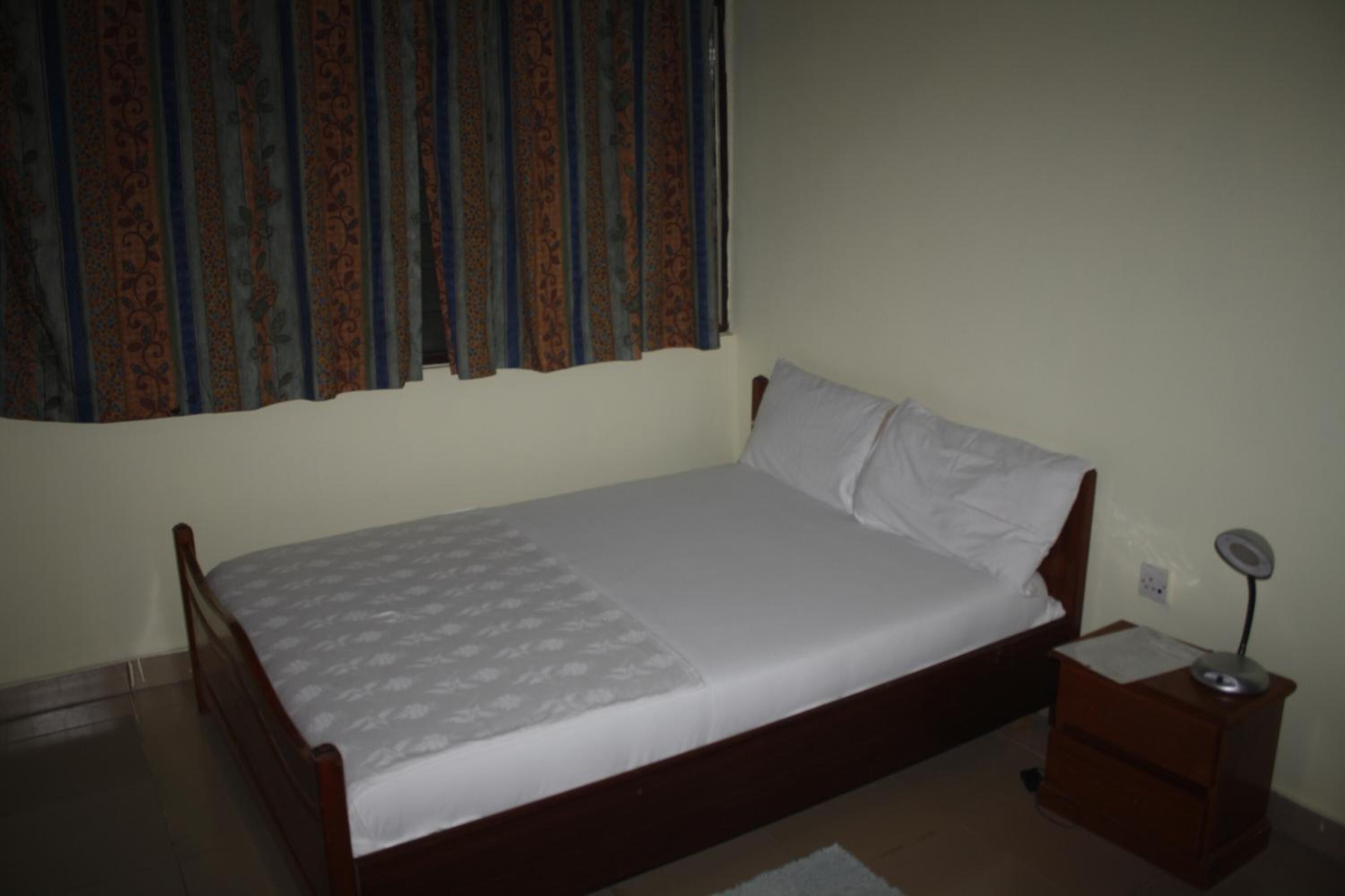 A single bed with a wooden frame, a white mattress, and a patterned bedspread. There is a nightstand with a lamp beside the bed.