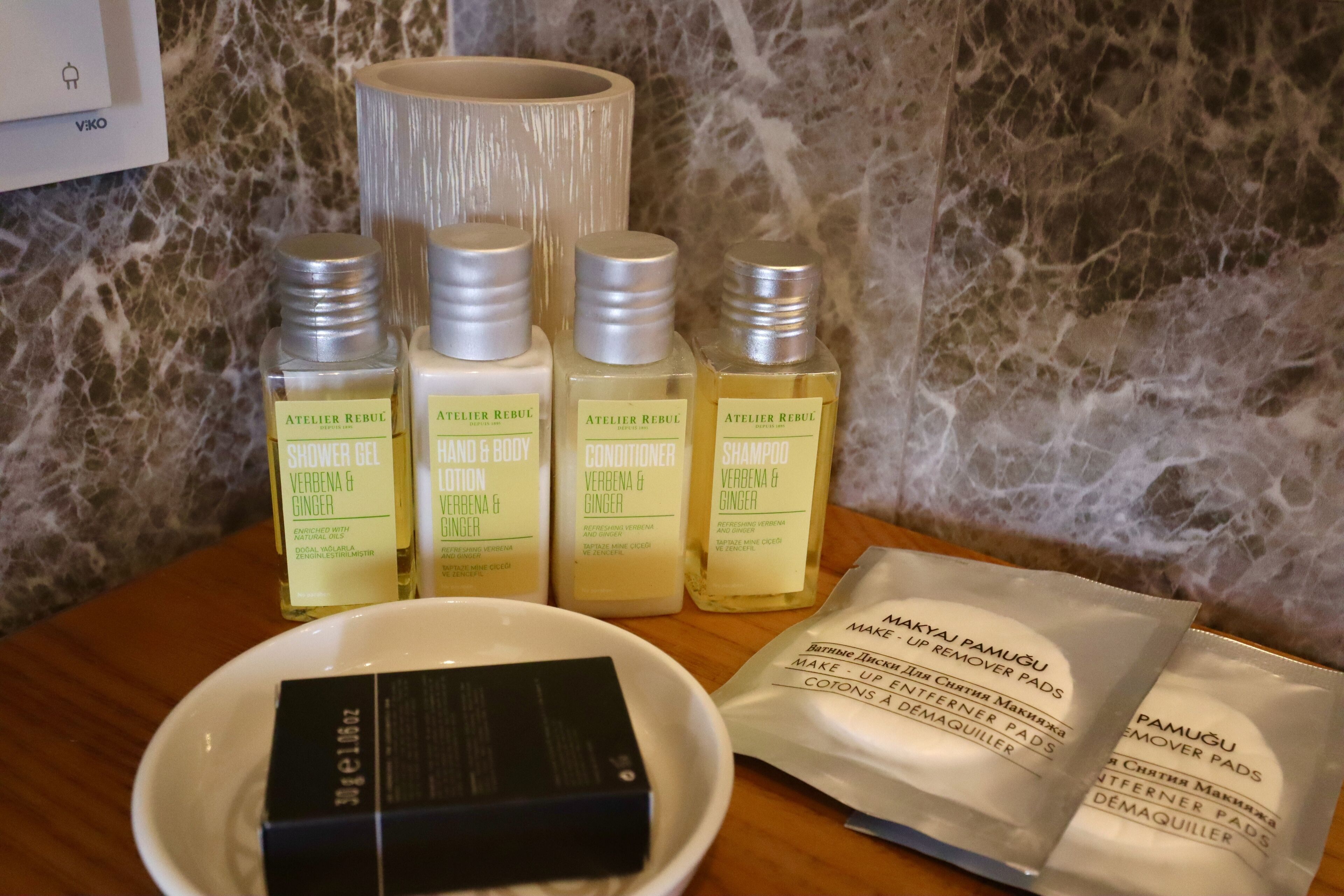 A bathroom with a marble wall, a white towel dispenser, and a set of toiletries including shower gel, body lotion, conditioner, and shampoo.