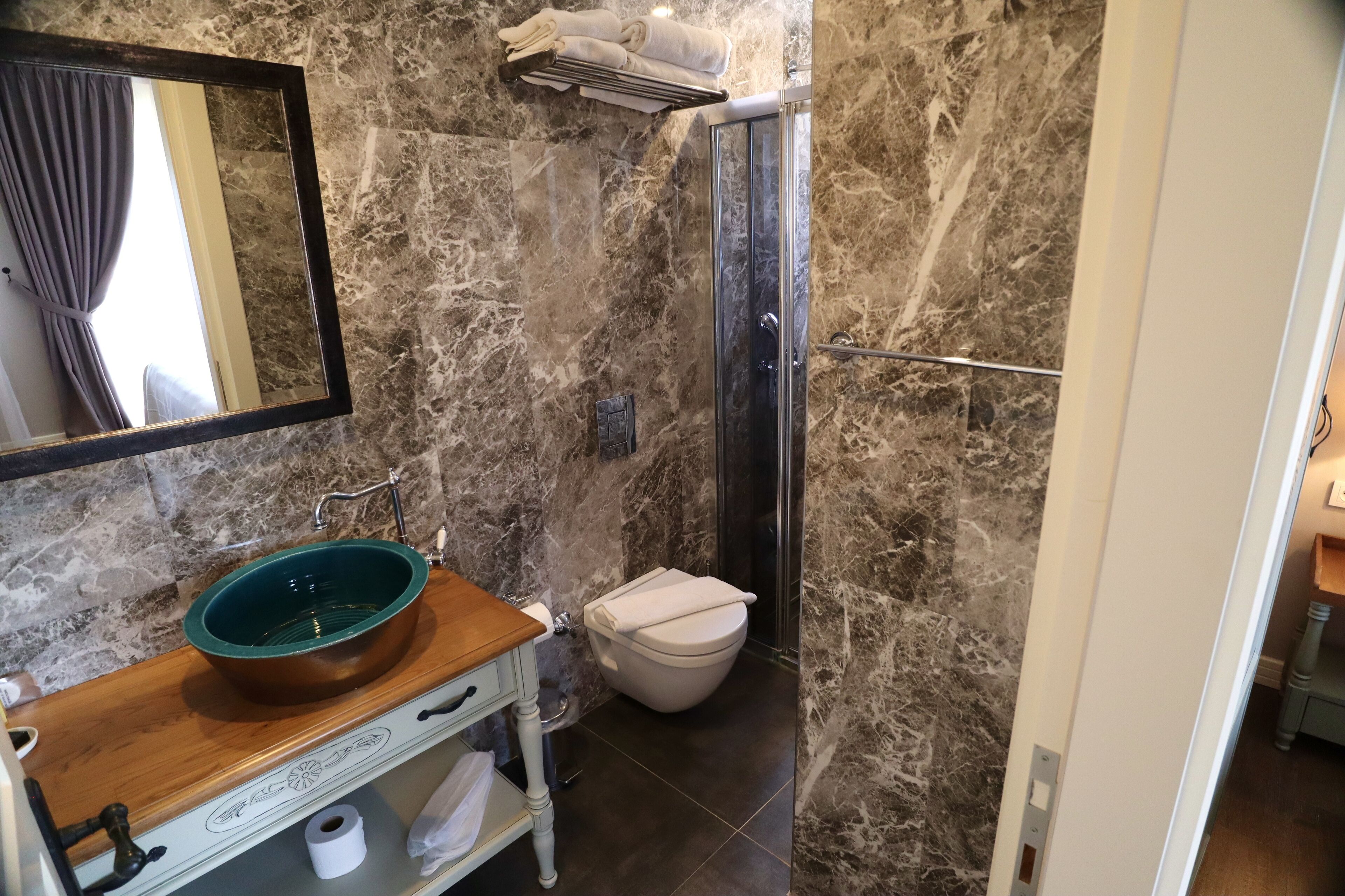 A bathroom with a marble wall, a wooden vanity with a bowl sink, a toilet, and a shower area.