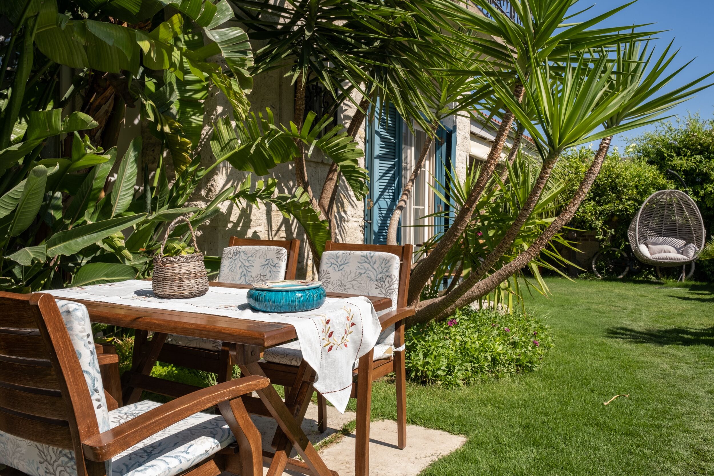 An outdoor dining area with a wooden table and chairs, surrounded by lush greenery and a palm tree.