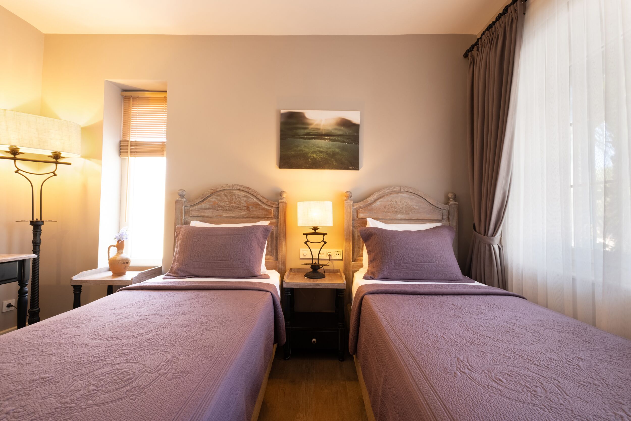A hotel room with two beds, a nightstand with a lamp, a painting on the wall, and a window with curtains.