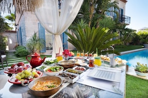 A table set with a variety of foods and drinks, a red teapot, and a menu in front of a poolside setting.