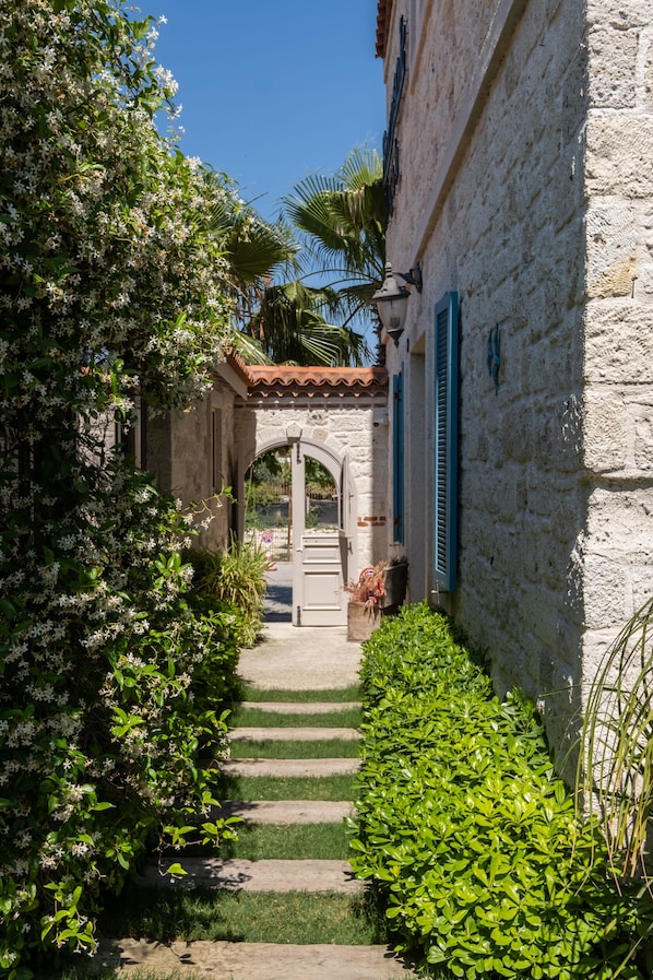 A stone building with a tiled roof, a narrow pathway leading to an archway, and lush greenery on either side.