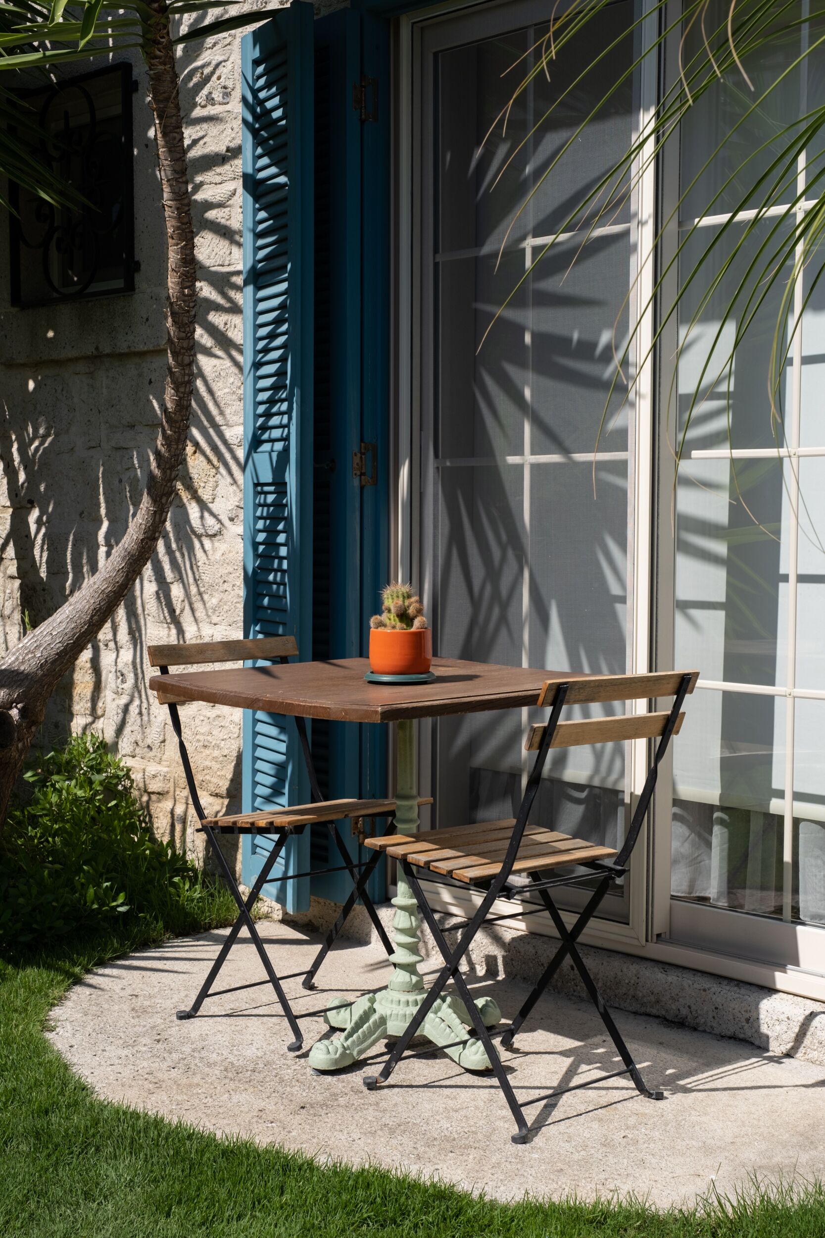 A patio with a wooden table and chairs, a potted plant, and a view of a building with blue shutters.
