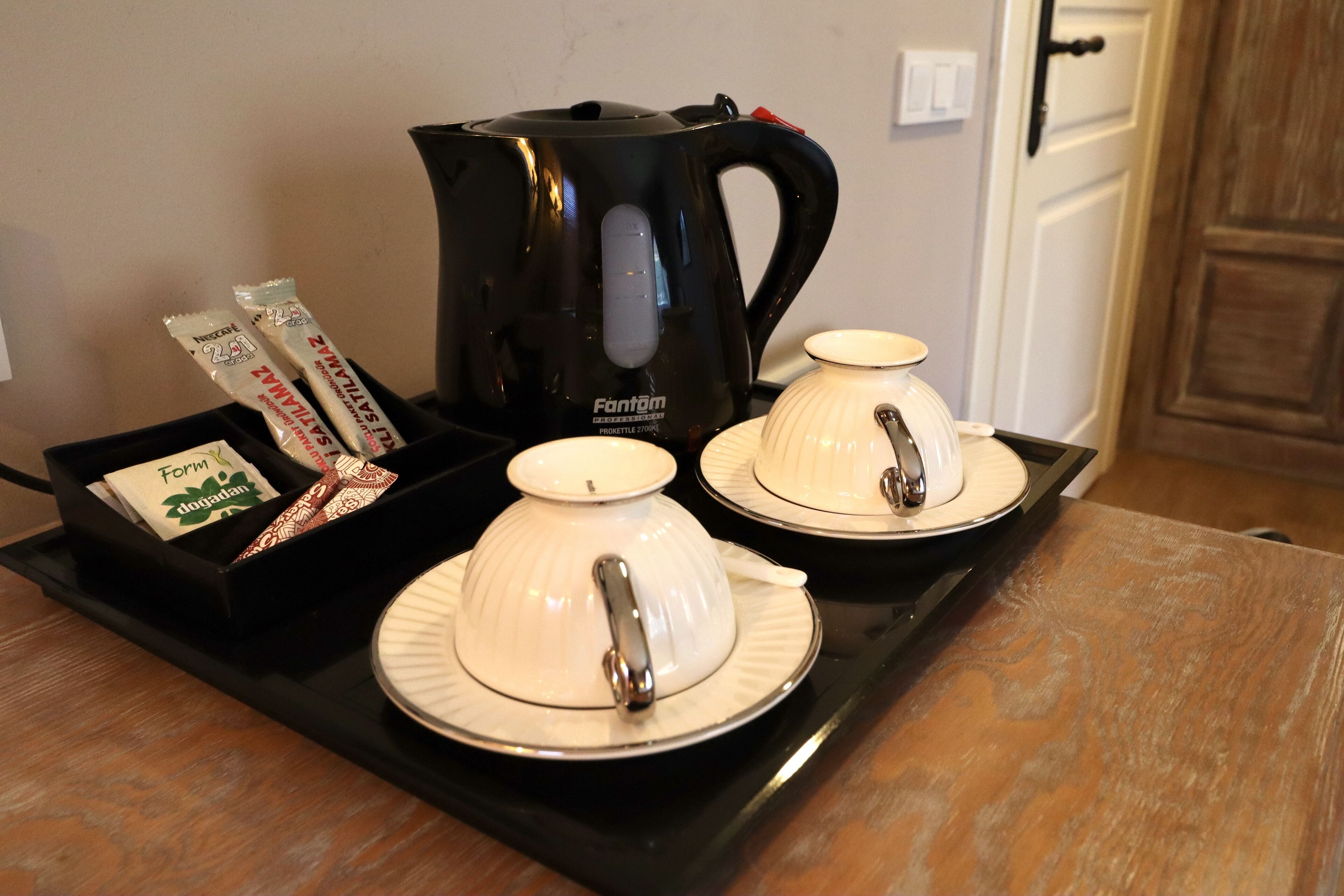 A kitchen counter with a black tray holding two white teapots with handles, a black electric kettle, and packets of snacks.