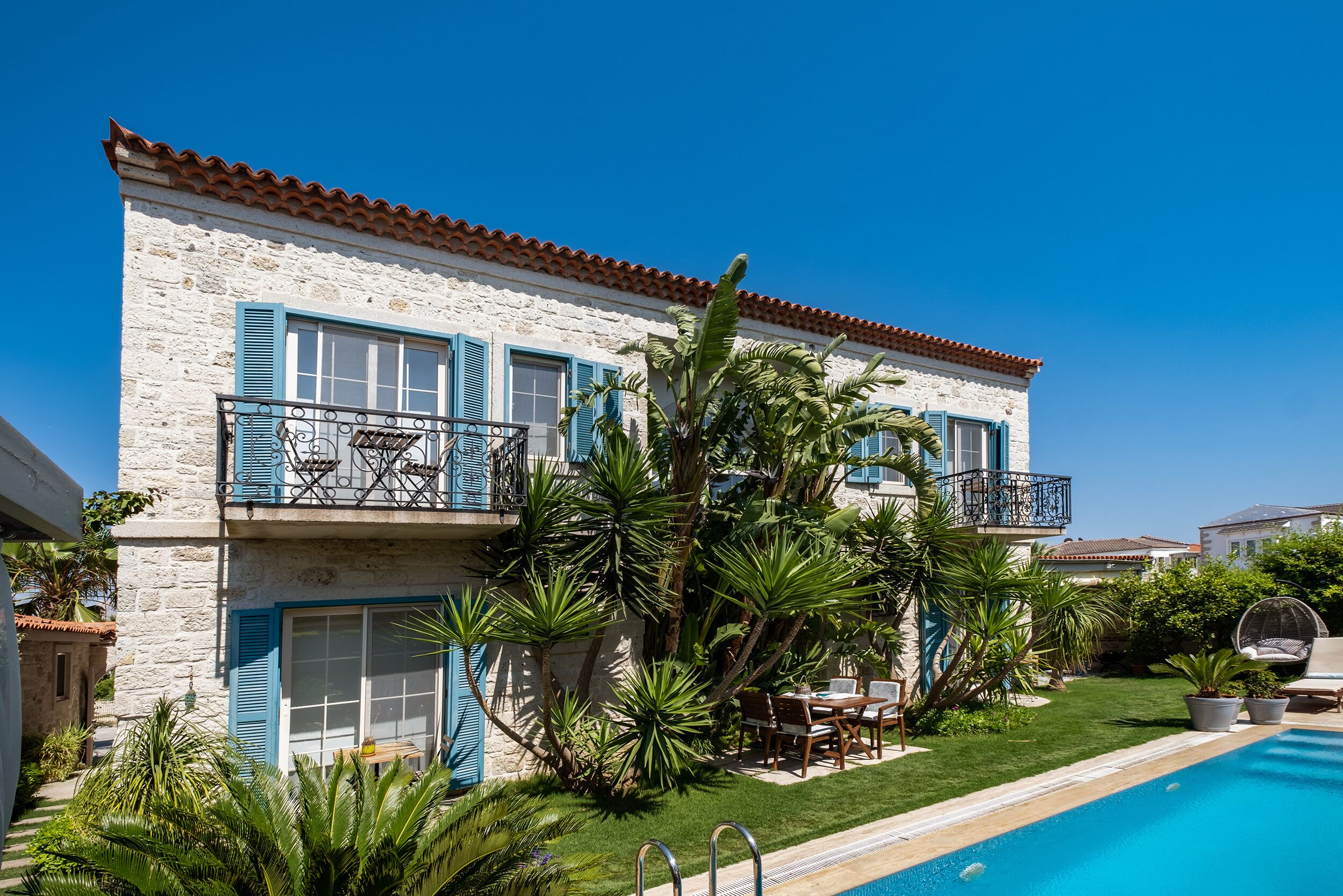 Un edifici de dues plantes amb piscina, un balcó amb persianes blaves i vegetació abundant.