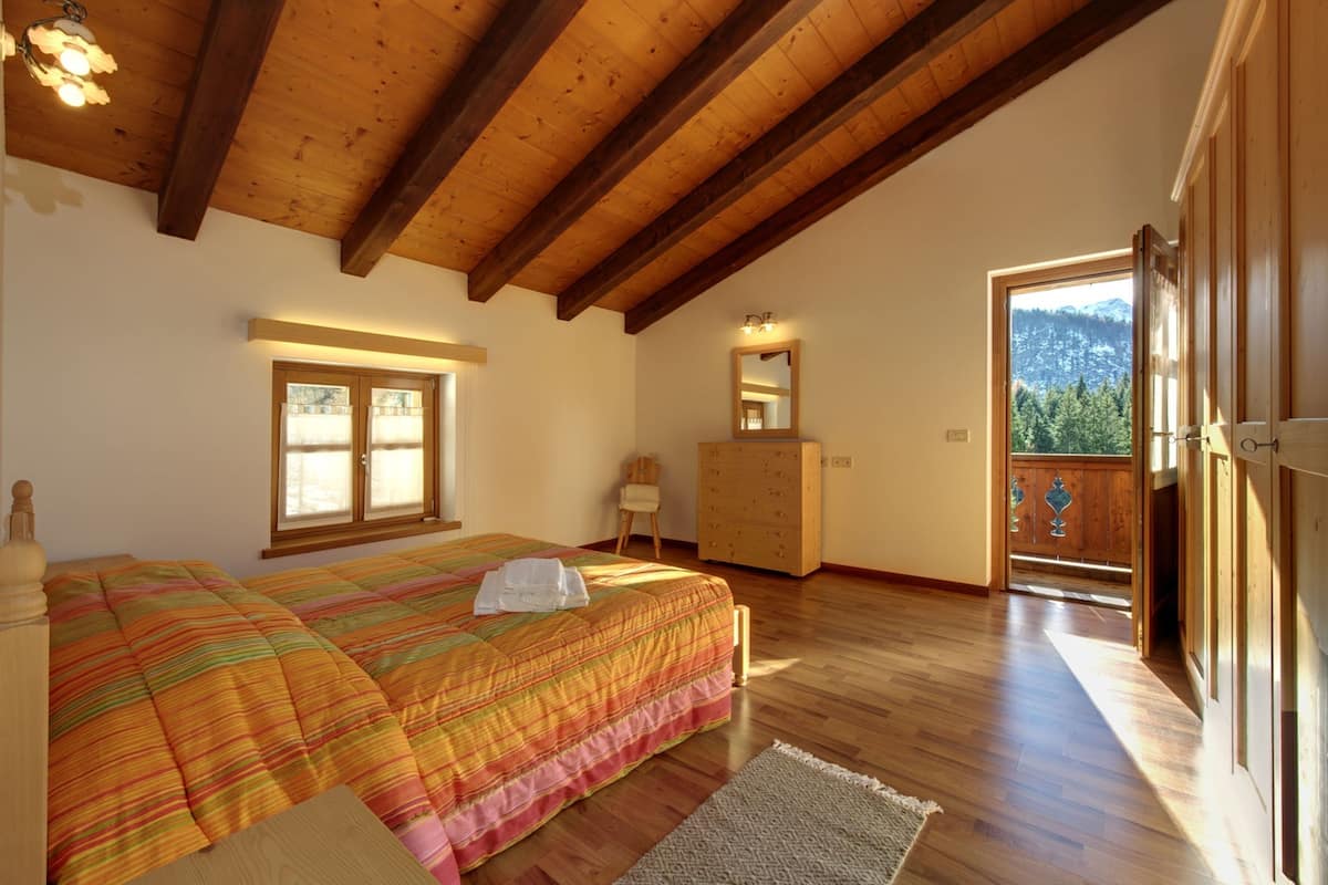 A bedroom with a wooden ceiling, a bed with a colorful bedspread, a dresser, a mirror, and a view of a mountain through a door.