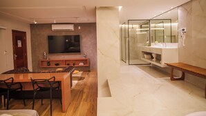 A modern living room with a TV, seating area, and a glass-enclosed bathroom with a vanity and bench. - W4 Hotel (Irece)