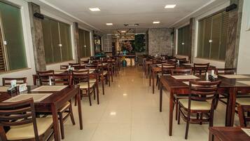 A spacious dining area with multiple wooden tables and chairs, a central counter, and large windows with green blinds.