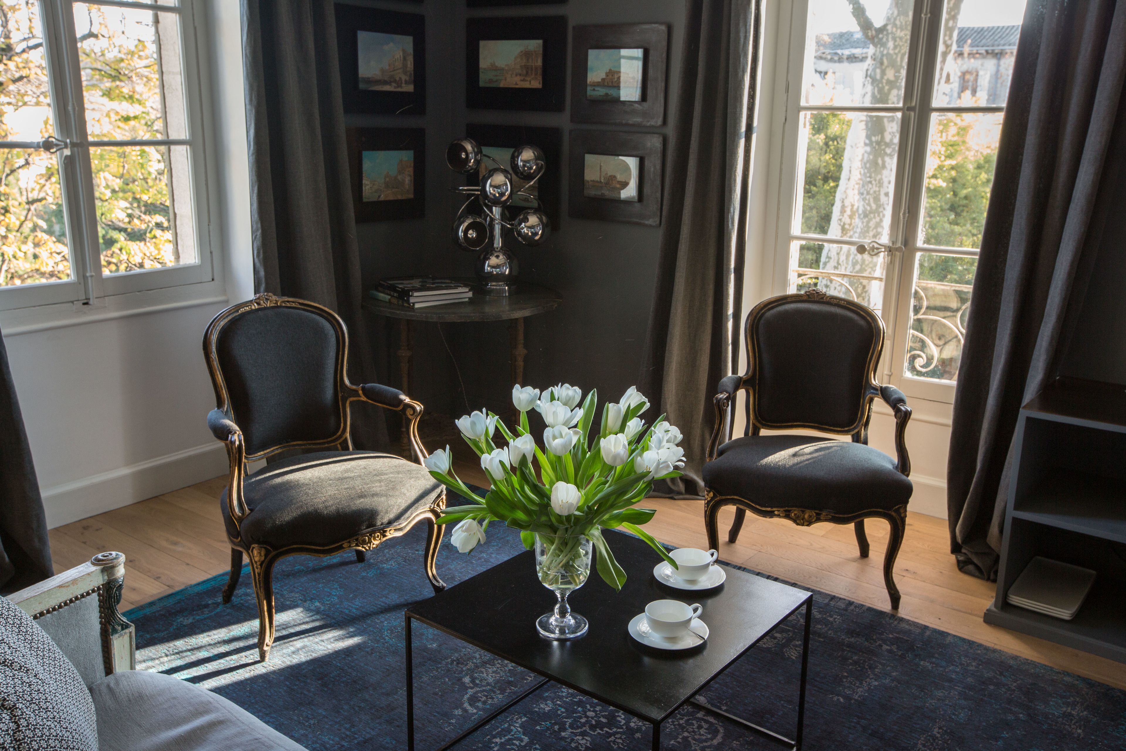 A room with two black upholstered chairs, a small table with a vase of white flowers, and a view of buildings through large windows.