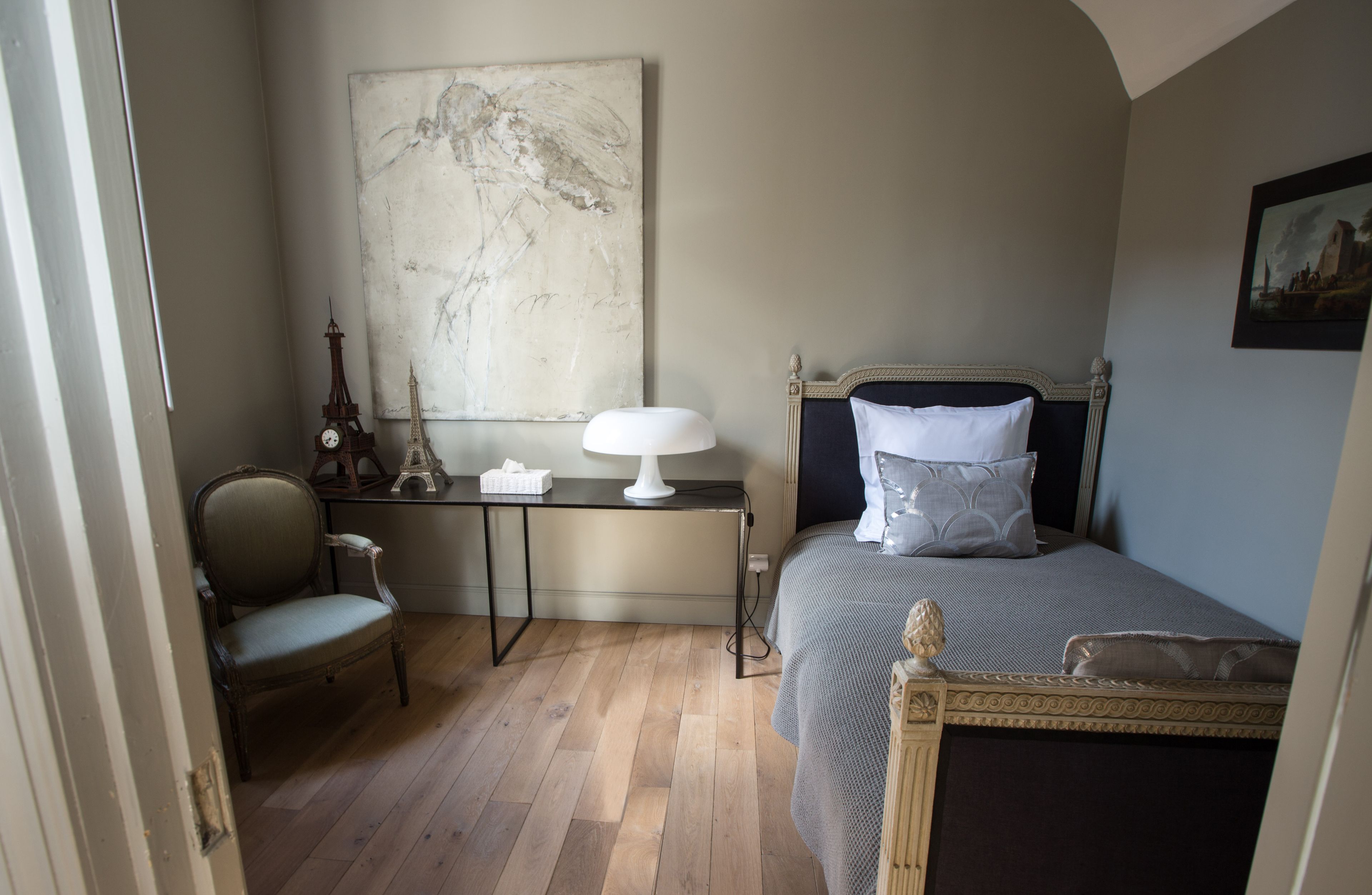 A bedroom with a bed, a chair, a desk, and a large painting on the wall.