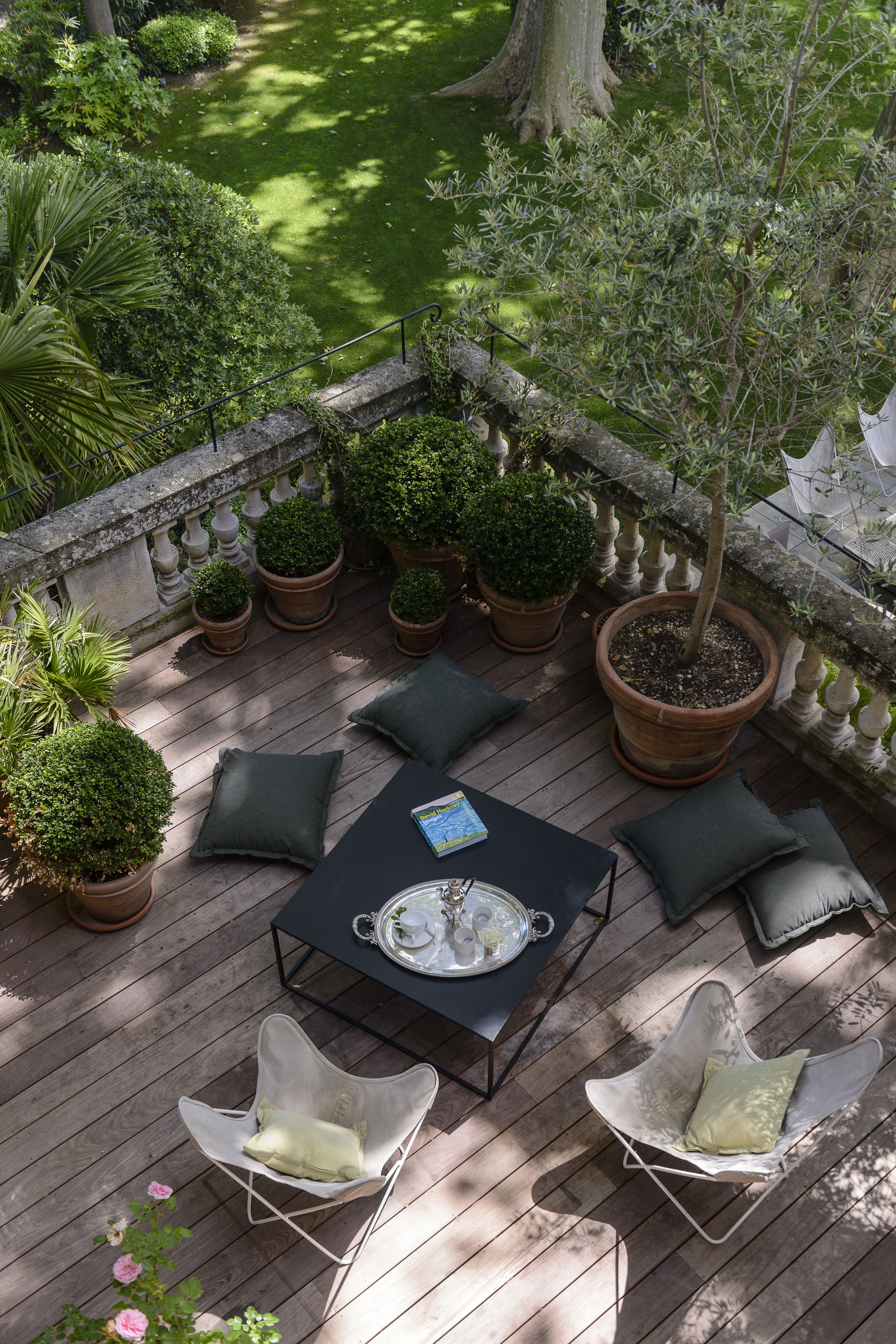 Um deck de madeira com mesa e cadeiras, cercado por plantas em vasos e vista para um jardim.