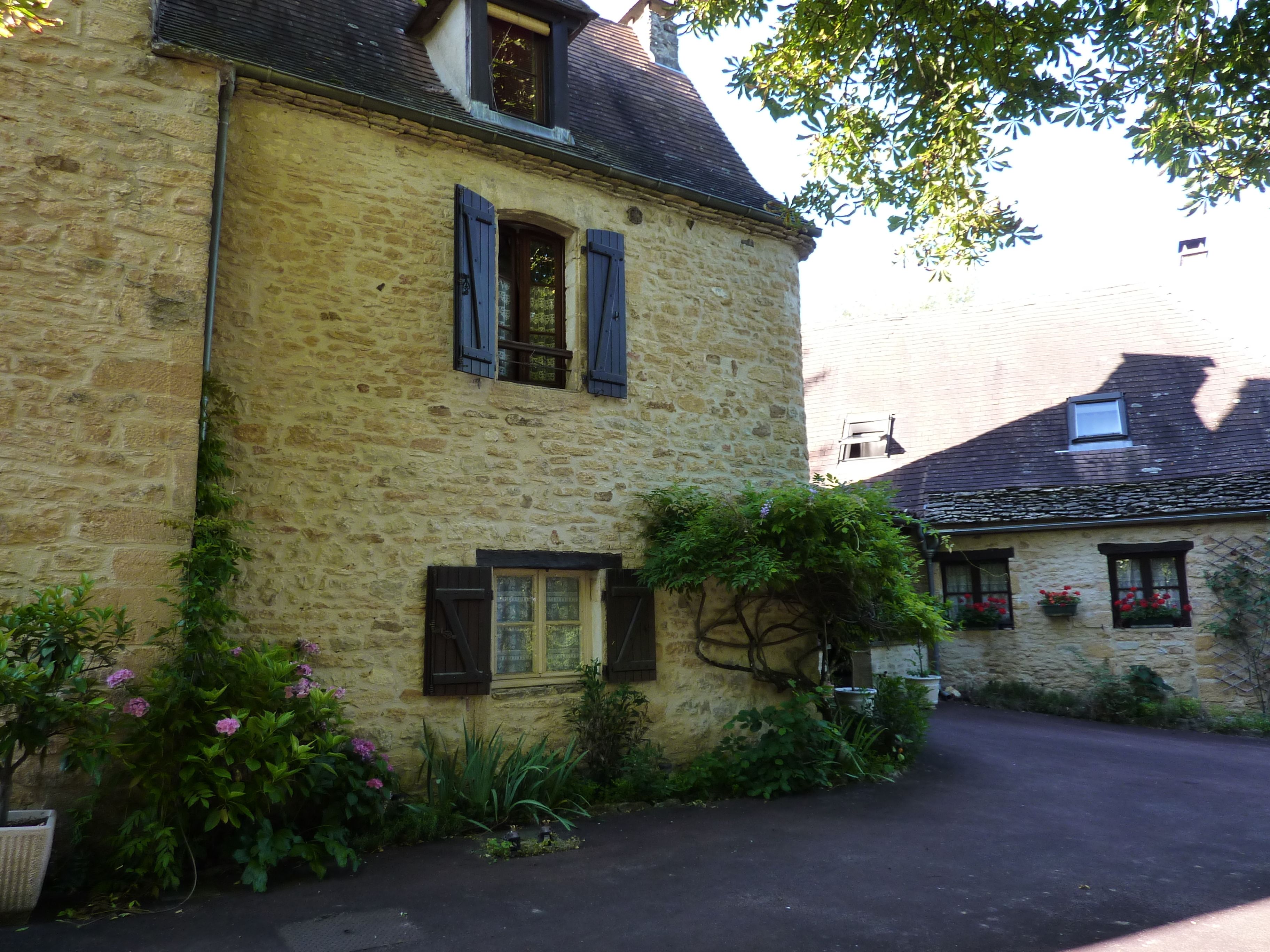 A stone building with a sloped roof, two windows with shutters, and a garden with flowers and plants.