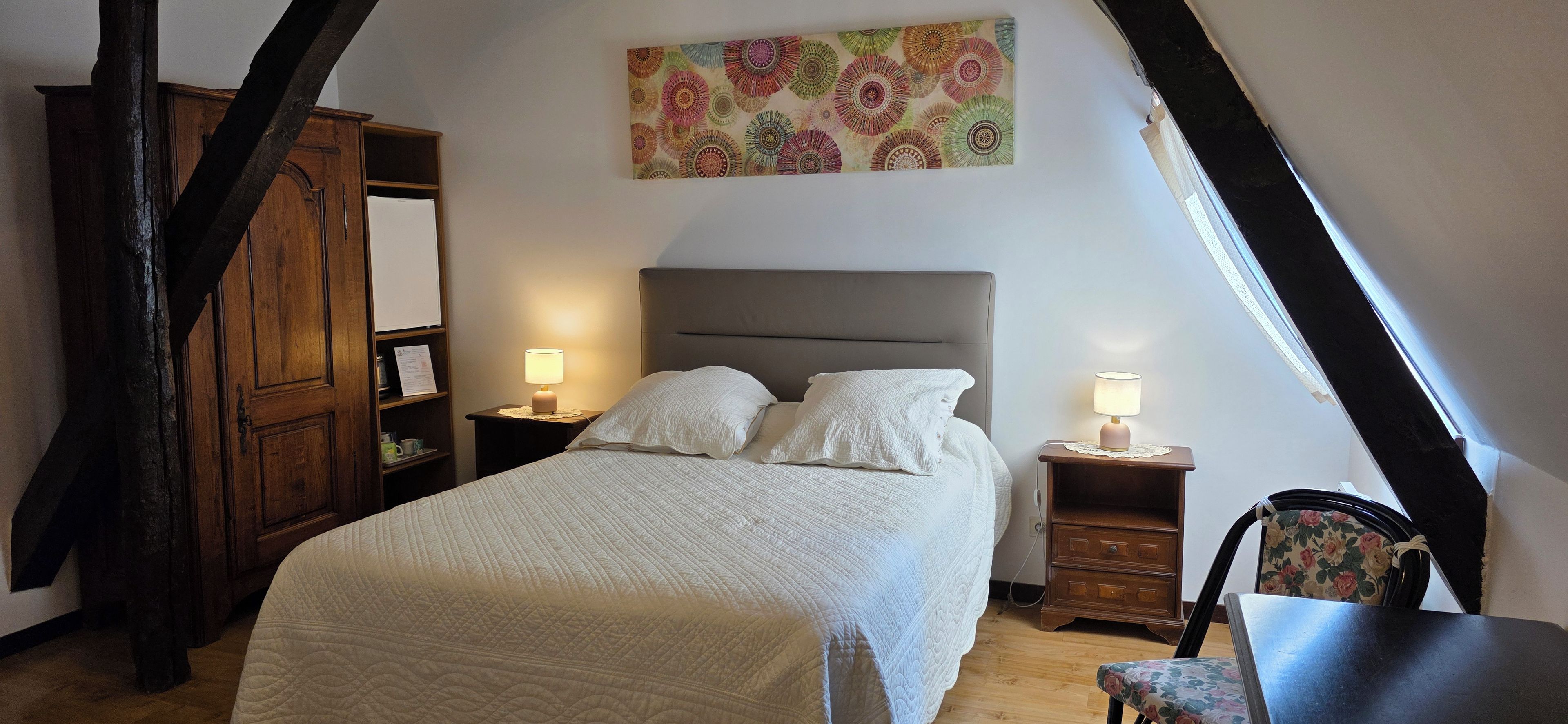 A bedroom with a bed, two bedside tables with lamps, a wooden wardrobe, and a floral artwork on the wall.