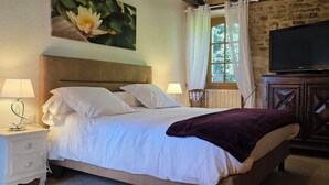 Een bed met witte lakens, een paarse deken, een televisie op een houten kast en een schilderij van een bloem aan de muur.