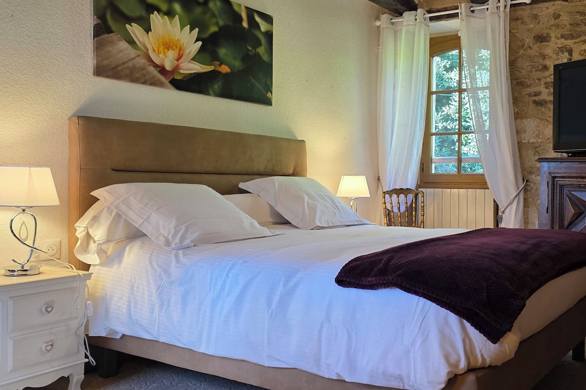 Een bed met witte lakens, een paarse deken, een televisie op een houten kast en een schilderij van een bloem aan de muur.